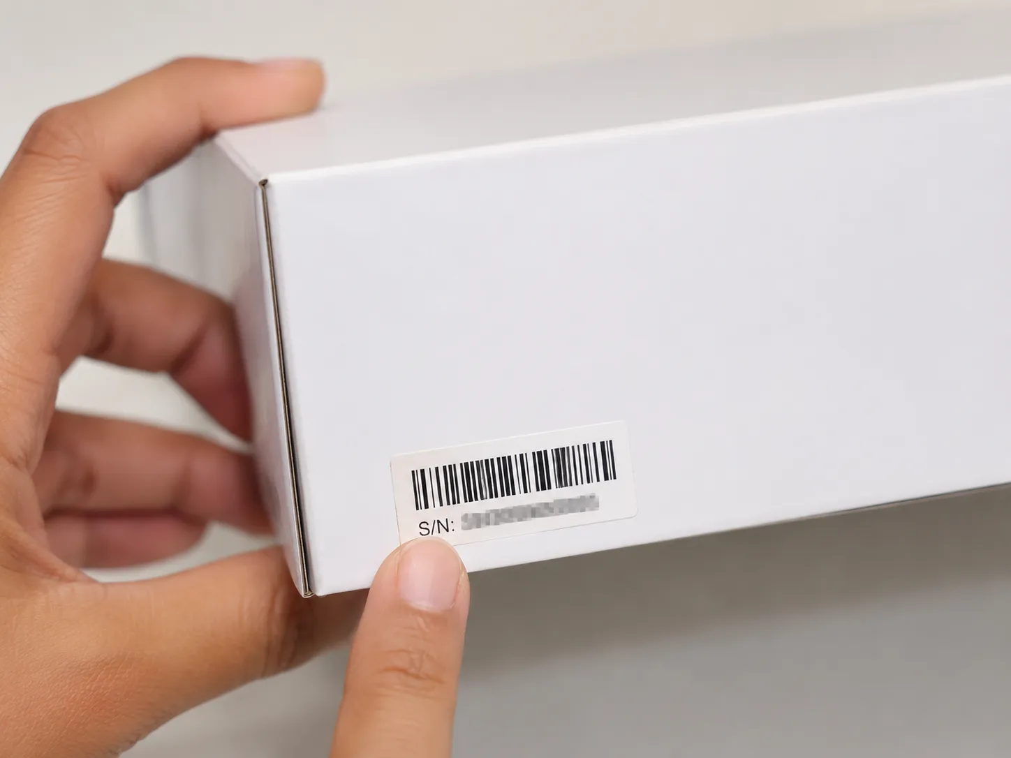 Hand pointing to the serial-number barcode label on the corner of a plain laptop box, illustrating where to find the serial number on retail packaging