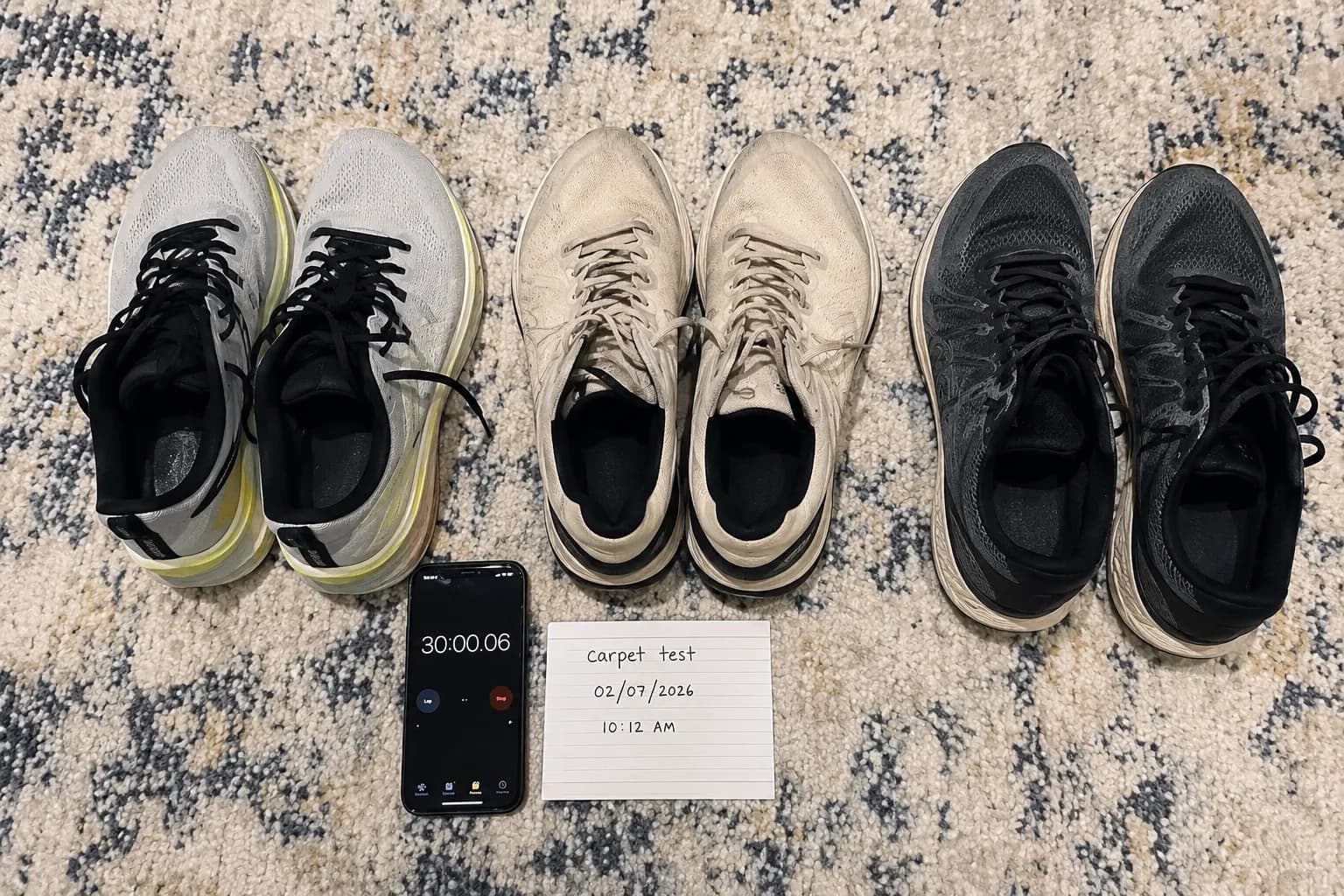 Overhead photo log showing three pairs of running shoes lined up on a patterned carpet beside a phone timer and a handwritten "carpet test" note with a timestamp, documenting an indoor wear trial.