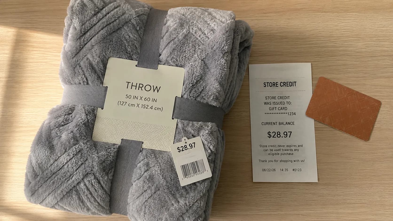Overhead flat-lay of a folded grey throw blanket with its price tag beside a store-credit slip showing a $28.97 balance and a generic gift card on a light wood surface.