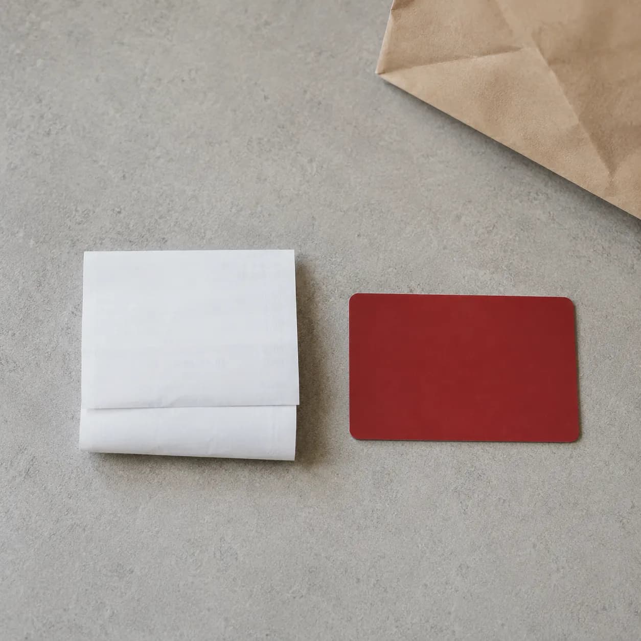 Overhead documentary-style view of a folded gift receipt beside a plain store gift card on a light gray stone surface, illustrating a return that was declined one day and accepted the following.