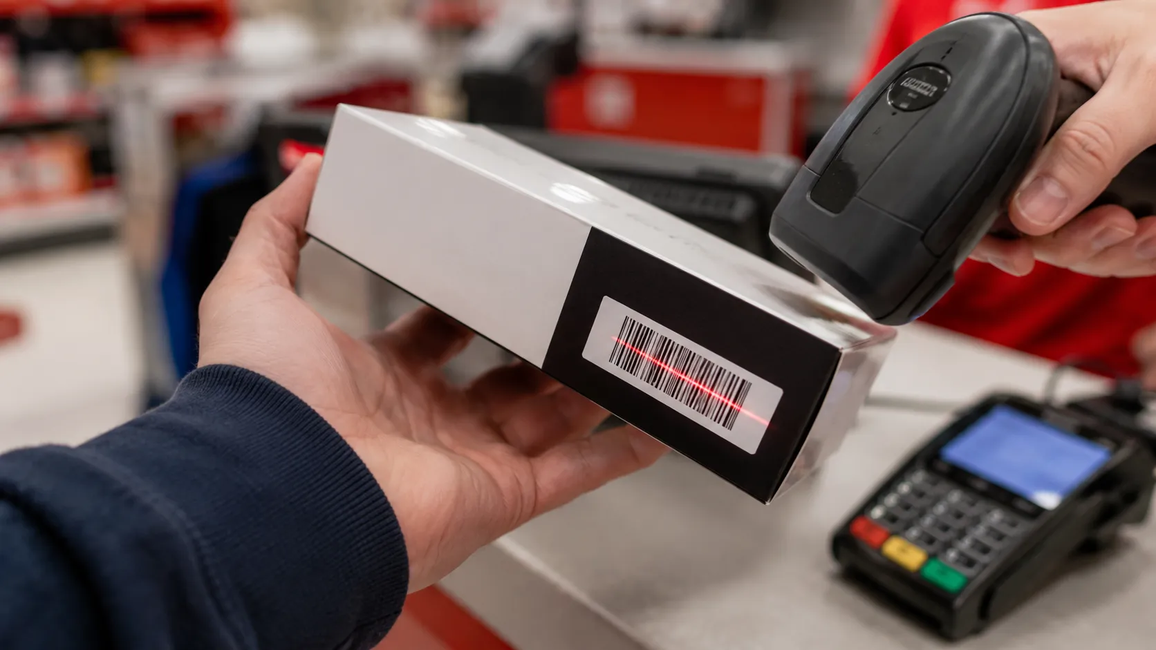 Close-up of a handheld retail scanner reading the serial barcode on a plain electronics box held by a customer at a big-box store returns counter, with a blurred service desk and card reader.